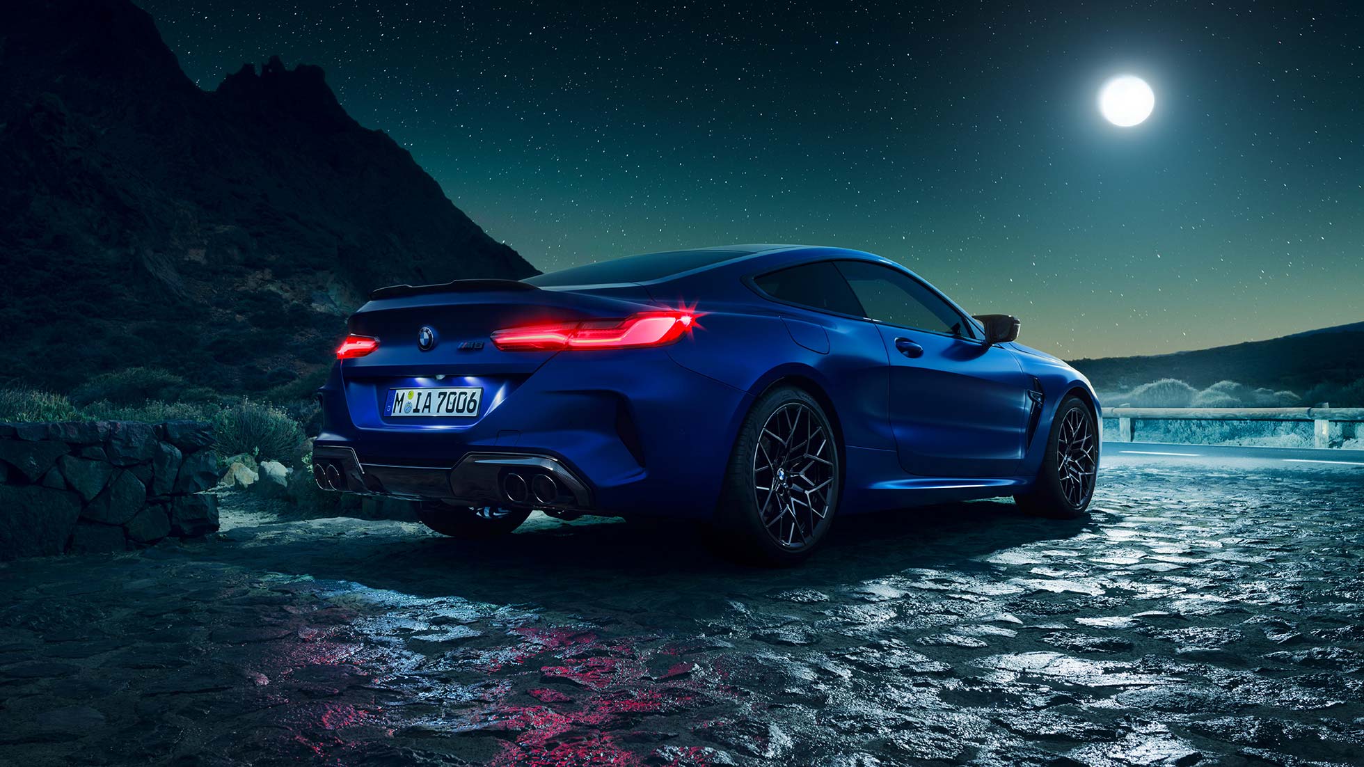 BMW M8 Coupé, three-quarter rear view. BMW M8 Coupé in Marina Bay Blue metallic with illuminated LED rear lights, standing three-quarter rear view with full moon.